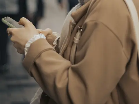 A person in a camel coat holding a phone in both hands