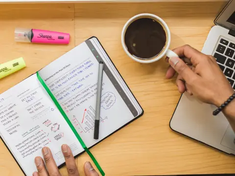 A notebook of plans, a coffee and a laptop on a wooden desk