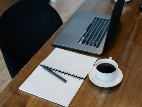 An open notebook, a pen, a coffee and a laptop on a wooden table