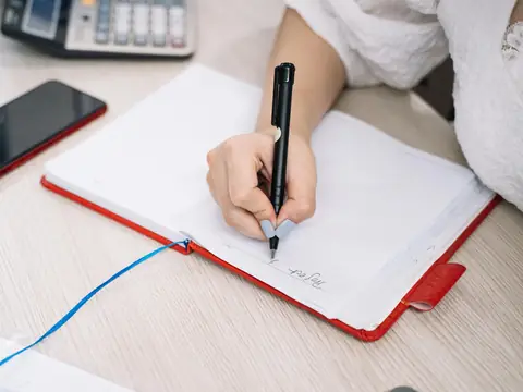 A hand writing in a notebook with a phone on the desk