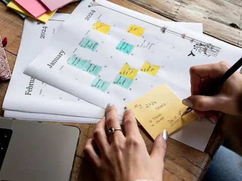 A hand writing on a sticky note beside a paper calendar