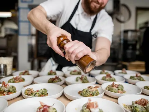 A cook seasoning rows of plated salads in a restaurant kitchen